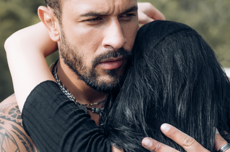 A man with tattoos hugs a woman with long black hair, both showing serious expressions.