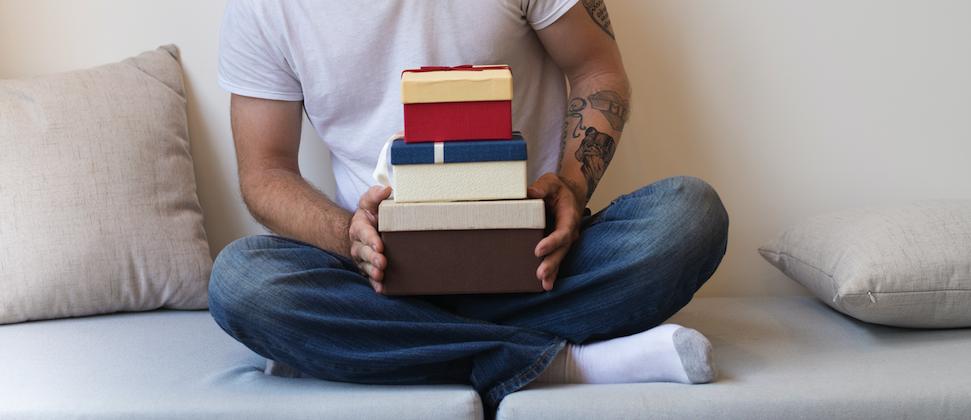 Person sitting cross-legged on a couch holding several stacked gift boxes, wearing a white shirt and jeans.