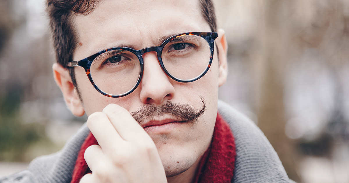 Man with glasses and a curled mustache looking at the camera, touching his face, wearing a scarf and coat.
