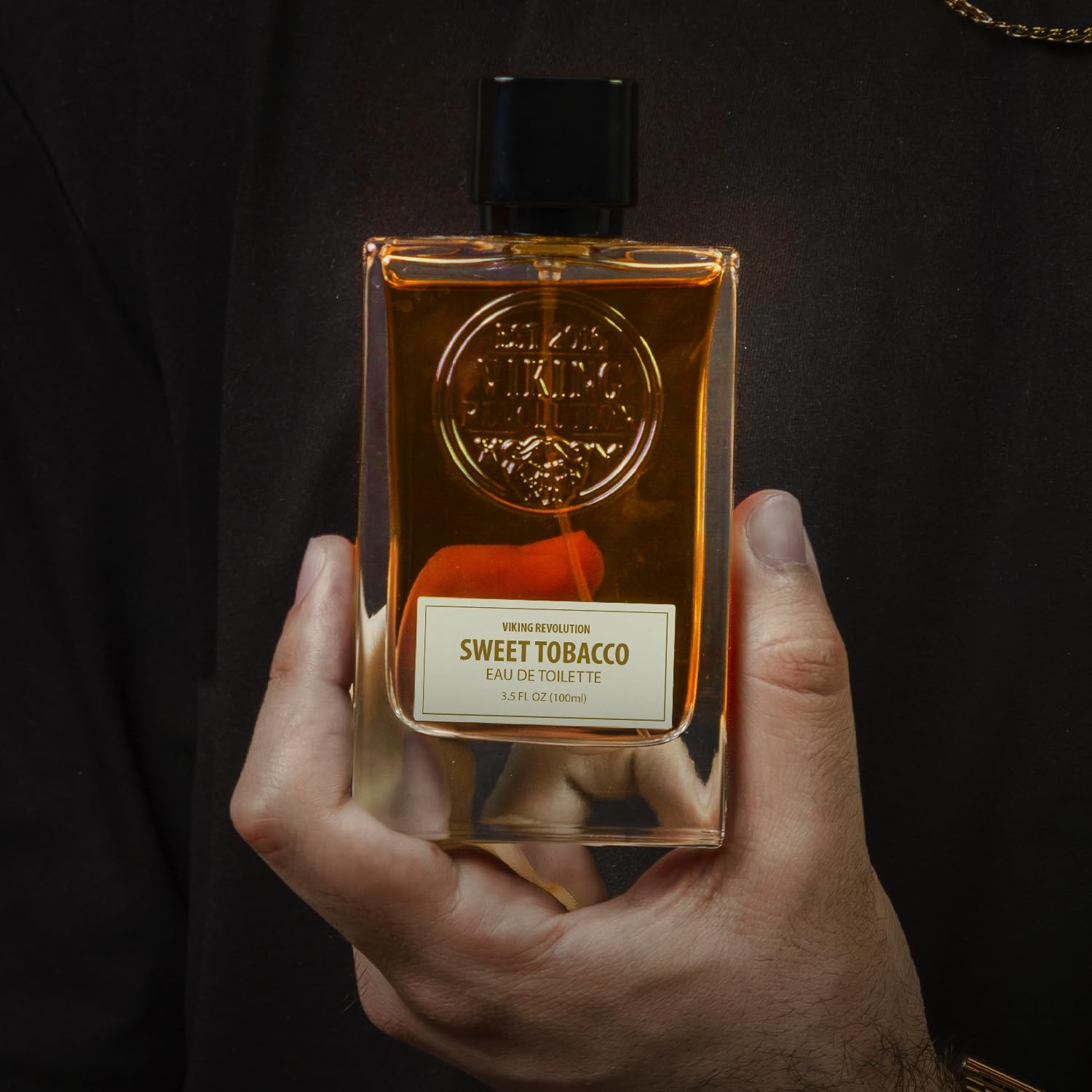 A person holds a bottle of Cologne for Men 3.5 fl oz from Viking Revolution against a dark background, evoking an air of mystery with its sweet tobacco notes reminiscent of Men's Cologne.