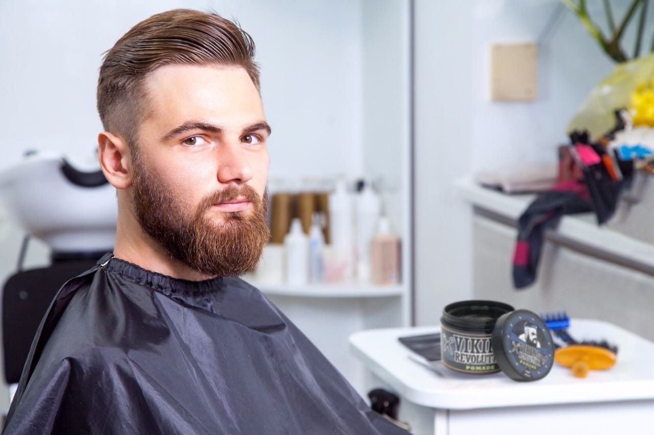 A bearded man with a fresh haircut sits in the barbershop, wearing a black cape and surrounded by Hair Pomade - 4oz. His confident smile reflects the satisfaction guarantee of exceptional hair grooming using natural ingredients.