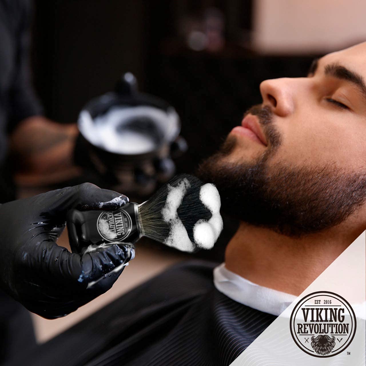 A man indulges in a luxurious beard treatment at the barber shop, where a Badger Hair Shaving Brush expertly applies the shaving cream for the perfect wet shave experience.