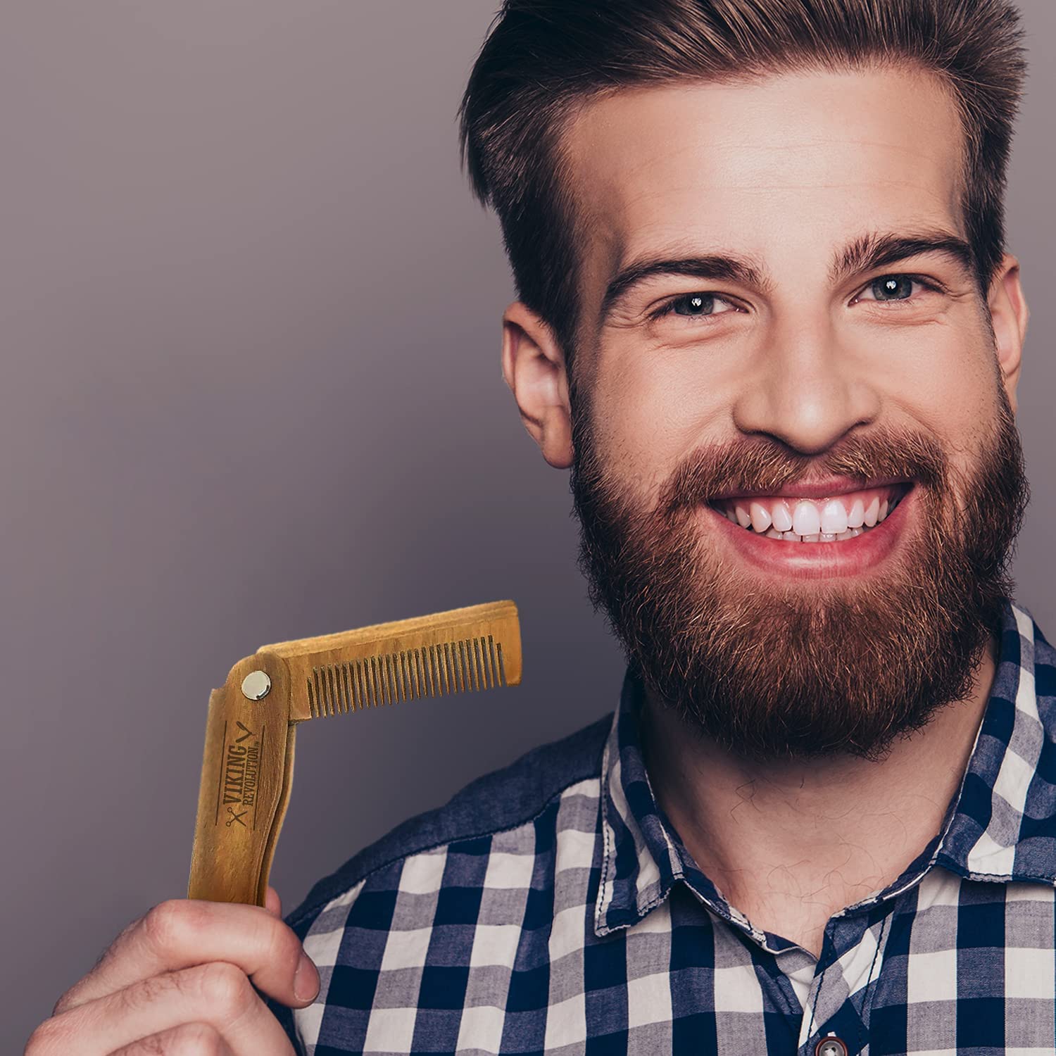 A confident man with a beard and checkered shirt smiles while holding the Folding Beard Comb w/Carrying Pouch against a gray background, showcasing his beard grooming routine.