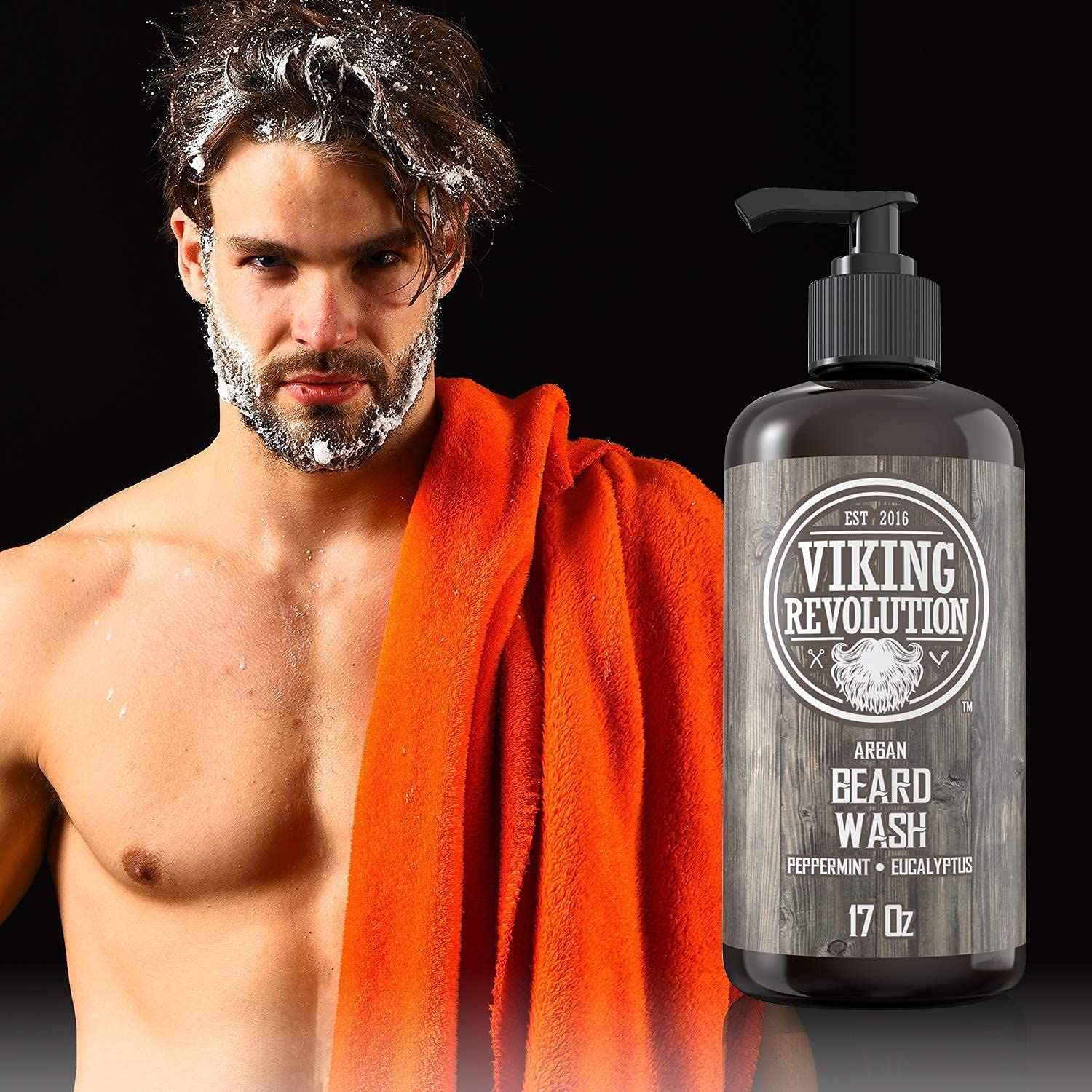 A man with facial hair, holding an orange towel, stands beside a large bottle of Peppermint & Eucalyptus Beard Wash 17oz, ready to enhance hair growth.