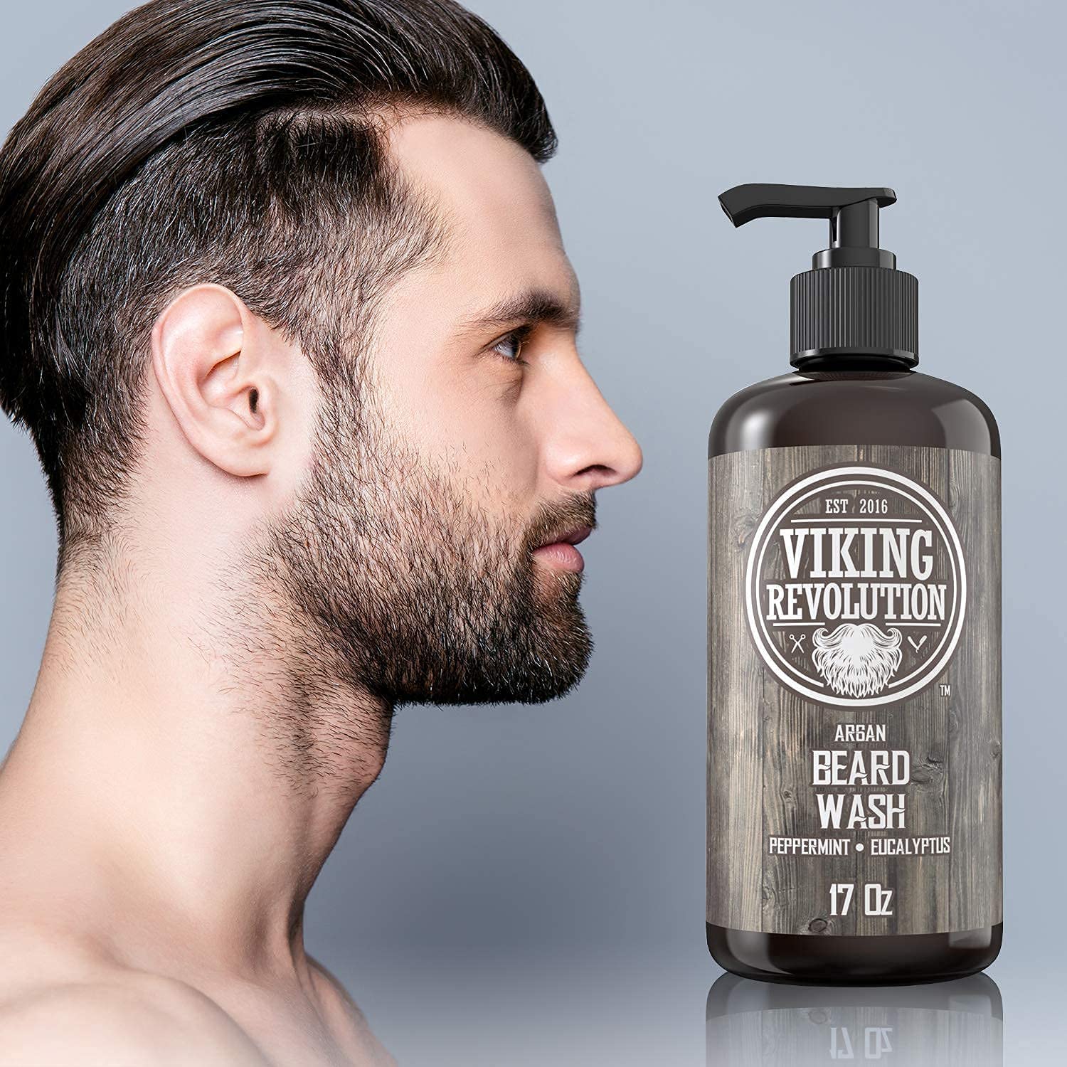 A man with a beard gazes thoughtfully at a Peppermint & Eucalyptus Beard Wash 17oz bottle on a gray background, contemplating its promise of enhanced facial hair growth.