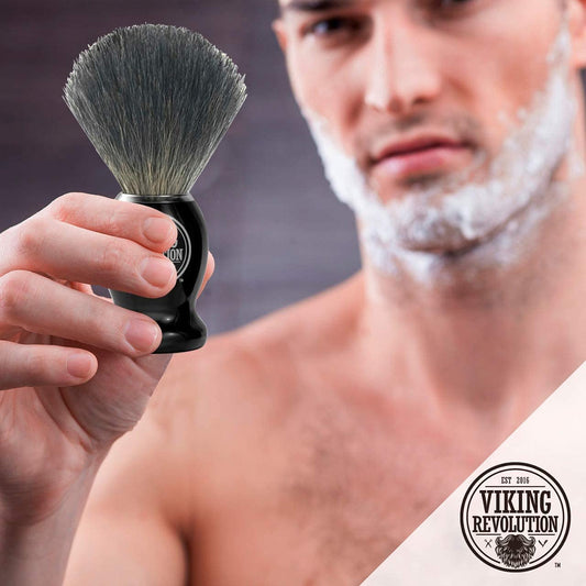 A man expertly handles a Badger Hair Shaving Brush, his face partially covered in rich shaving cream, showcasing the Viking Revolution logo as he enjoys a traditional wet shave.