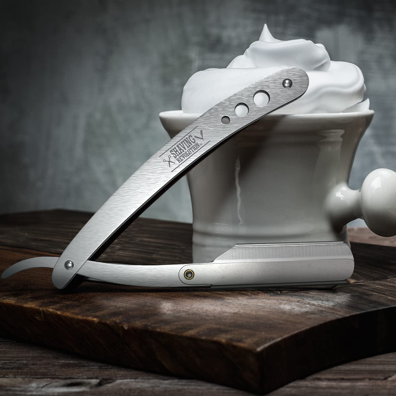 A Straight Edge Barber Razor with 100 single edge blades rests on a wooden surface, next to a mug filled with shaving cream made from premium ingredients.