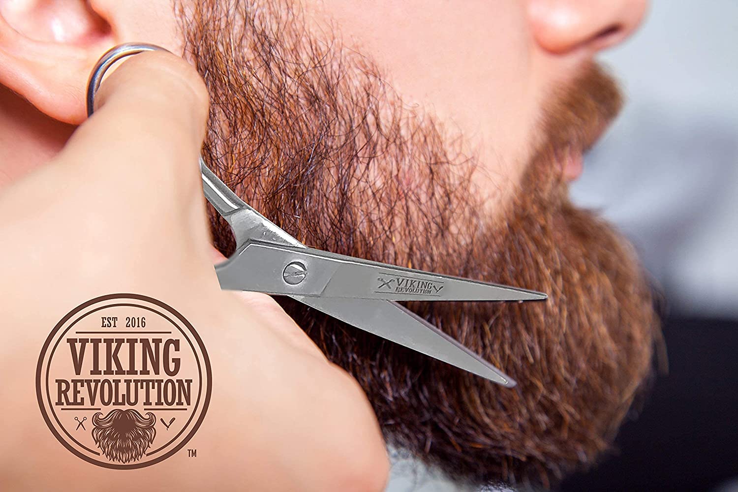 Close-up of a person trimming a brown beard with Viking Revolution scissors; logo in the corner.